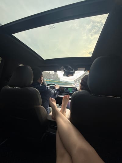 Inside a car, a driver steers with one hand. A passenger has their bare feet on the dashboard near the navigation screen. A second passenger's bare legs are crossed on the center console. The car has a sunroof through which a cloudy sky is visible. The interior is dark, with black leather seats.