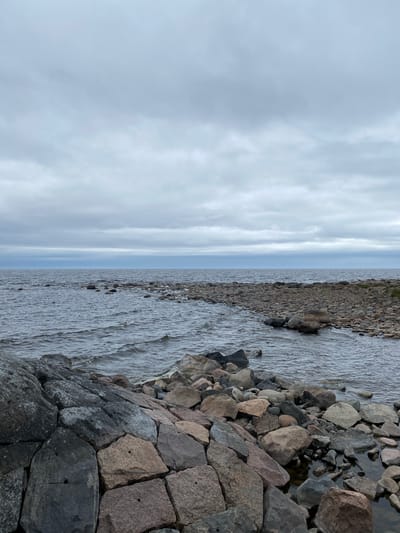 The image depicts a rocky shoreline on an overcast day. The foreground features a jumble of large, irregularly shaped rocks in various shades of gray and brown, some with visible drill holes. These rocks transition to a more densely packed, smaller pebble-like rocky shore extending into the water. The water itself is dark and appears to have small waves. In the middle ground, the water stretches out to a distant horizon line, where it meets a pale blue-gray sky filled with layers of clouds. The overall scene is a natural landscape with no signs of human activity or man-made structures. The location is identified as a rocky shore in Моторное, Russia.