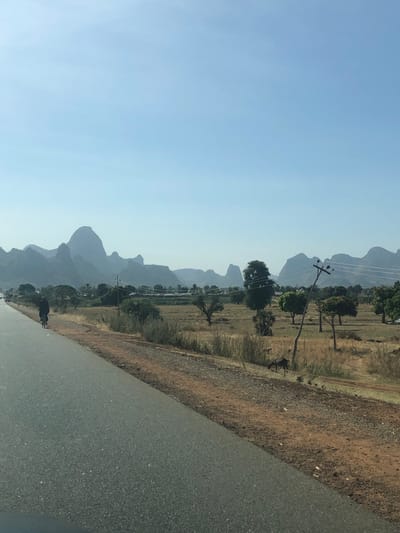 This image presents a wide-angle view of a rural landscape within Dott, Nigeria, under a clear, pale blue sky. A two-lane asphalt road extends diagonally from the lower left toward the upper left. A single individual, dressed in dark attire, is observed cycling a bicycle away from the viewer on the left side of the road. To the immediate right of the road, an unpaved reddish-brown dirt shoulder transitions into an expansive field. This field contains dry, light-colored vegetation interspersed with scattered green trees. A wooden utility pole, visibly leaning to the right, is positioned within this field, featuring overhead wires. Beneath the utility pole, a dark-colored animal, consistent with a goat, is grazing. The background is dominated by a distinct range of irregularly shaped, rounded mountains rising against the horizon.