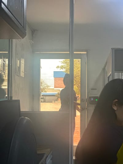 The image depicts an indoor scene, likely an office or administrative building, with a person silhouetted in the doorway leading outside. The doorway reveals an outdoor environment bathed in bright daylight, with trees and buildings visible. The interior is dimly lit and cluttered with office furniture, including chairs and what appears to be a computer monitor in the foreground. On the wall near the doorway, a security camera is visible. There is also a digital lock system mounted on the wall next to the door. A portion of a woman's face and hair is visible on the right side of the frame, suggesting she is seated inside. The overall impression is one of an active but somewhat confined workspace. The location is identified as Juan Mina, Colombia.