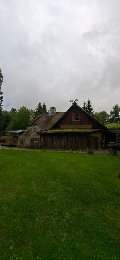 In Saula, Estonia, this image captures a rustic wooden building with a moss-covered roof and a stone chimney. Two decorative dragon-like figures adorn the peak of the steeply pitched roof. A wooden sign with yellow lettering, partially obscured by greenery, is mounted above a lower section of the building. The structure is surrounded by a wooden fence and a well-maintained green lawn. Mature trees form a backdrop under a cloudy sky. The scene depicts an outdoor setting with historical or traditional architecture.