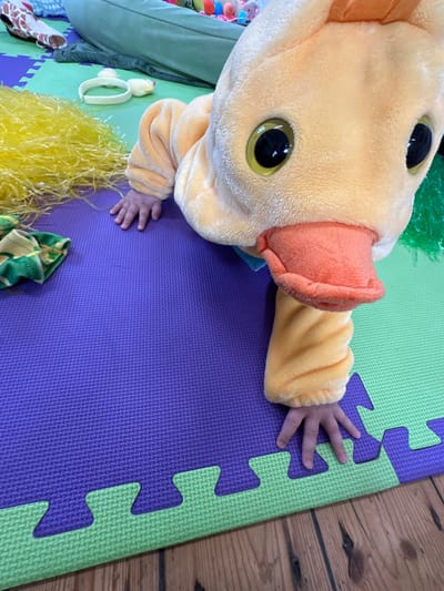 A young individual, wearing a plush yellow duck costume with prominent black and yellow eyes and an orange beak, is positioned on interlocking foam play mats. The individual's bare hands are resting on the textured purple and lime green mats, consistent with a crawling or kneeling stance. To the upper left of the subject, a substantial pile of bright yellow shredded plastic material is present on the purple mat, alongside a folded piece of green and white patterned fabric. A white headband adorned with two small yellow chick figures is also visible near the yellow material. In the background, an olive-green beanbag chair and other blurred, colorful objects are partially visible, suggesting a play environment. The floor beneath the play mats consists of wooden planks. The scene is located in Prestbury, United Kingdom.
