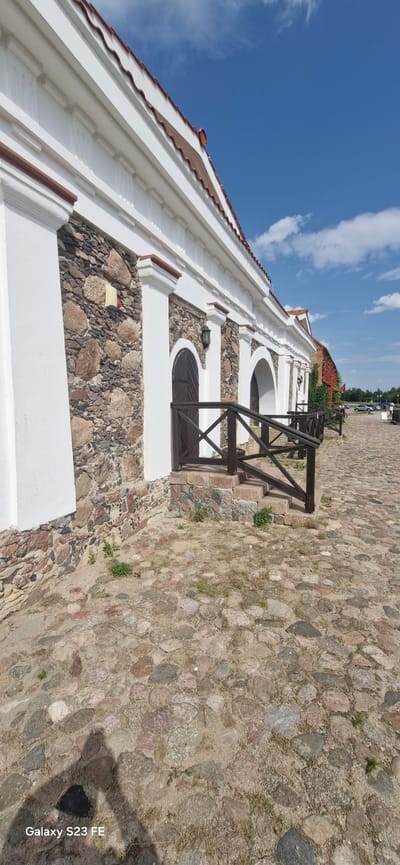 This is a ground-level, wide-angle photograph of the exterior of a historic building in Pakruojis, Lithuania. The building features a white facade with decorative cornices and arches, contrasting with its lower section, which is constructed of rough, uneven stone. Dark wooden doors are visible within some of the arched openings. A dark wooden railing with cross bracing leads up a short set of stone steps to one of the entrances. The ground in front of the building is paved with irregular cobblestones and patches of sand and sparse green vegetation. The sky above is bright blue with scattered white clouds. In the distance, a road with parked vehicles and trees can be seen. The image is captured on a sunny day.