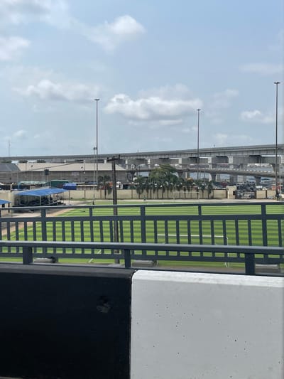 The image depicts an elevated view of an artificial turf football field located in Ebute-Metta, Nigeria, under a partly cloudy sky. In the immediate foreground, a black and white concrete barrier topped with a dark gray metal railing is visible. Beyond this barrier, the football field features alternating dark and light green stripes. Two white goalposts are discernible at the far end of the field. One small, dark human figure is observed walking towards the right, near the center of the field, while a second, smaller dark figure is positioned further left. A single light-colored football rests on the field to the left of the central figure.
Behind the field, a light-colored wall or fence runs horizontally, flanked by several palm trees. To the left, a structure covered by a blue tarp is partially visible. In the mid-distance, industrial-type buildings with various roof configurations are present, alongside multiple tall, slender light poles. The background is dominated by a complex network of multi-level concrete elevated bridges or overpasses, with vehicles moving on the uppermost structure. Faintly visible beneath these structures are what appear to be bodies of water and areas densely packed with vehicles and containers, indicative of a port or industrial environment.