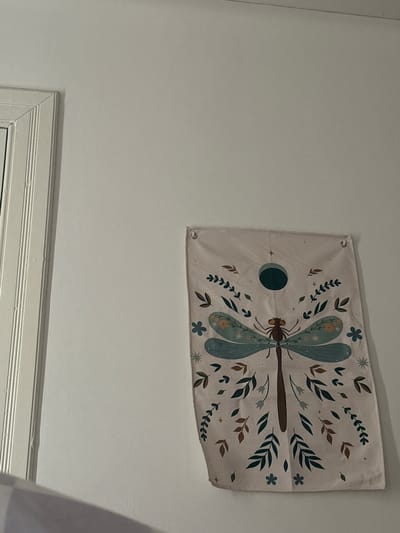 A rectangular tapestry featuring a dragonfly design hangs on a white wall. The dragonfly is surrounded by stylized leaves, flowers, and a dark green circle resembling a moon phase. Below the tapestry is a white window frame. The location is likely within a building in Aberdeen City, United Kingdom.
