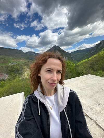 A photograph depicts a woman posing for a selfie with a mountainous landscape in the background. The woman has fair skin and reddish-brown curly hair and is wearing a white t-shirt, a light gray hoodie, and a black jacket. She is standing on a concrete platform or wall. The background features a valley with green hills and a mountain range under a partly cloudy sky. Based on the landscape, specifically the deep valley and mountain range, this image was likely taken in the area of Đurđevića Tara, Montenegro.