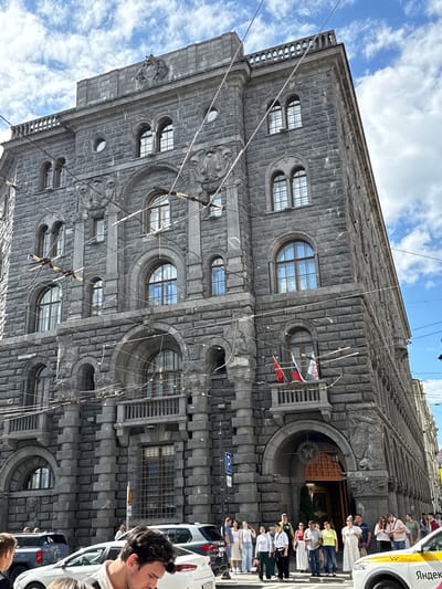 The image captures a street view of a grand, imposing stone building in Saint Petersburg, Russia. The architecture is characterized by heavy, rough-hewn grey stone blocks, arched windows, and decorative relief sculptures adorning the facade, including what appears to be a crest with a lion-like creature at the top. Wires, likely for trams or trolleybuses, crisscross the sky in front of the building.
On a balcony on the third floor, three flags are visible: one red, one white and red (likely Russian), and another white and red flag. Below the balcony, there's an ornate entrance to the building with carvings around the archway.
Several people are standing on the sidewalk in front of the building, some looking towards the entrance, others conversing. They are dressed in casual summer clothing. Several cars are parked or driving on the street, including a white taxi with "Яндекс" branding, indicating it's part of the Yandex.Taxi service, a common ride-hailing platform in Russia. The sky is partly cloudy with blue sky visible. The overall impression is of an urban setting on a bright day, highlighting a significant historical building.