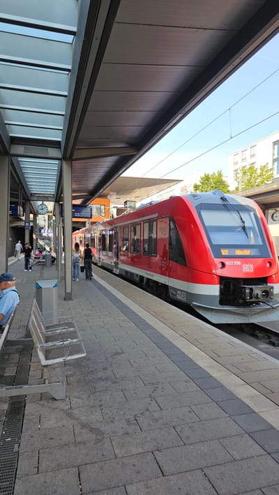 This is a photograph of a train station platform in Neu-Ulm, Germany. A bright red and silver train is parked at the platform, with its doors closed. The platform is paved with grey tiles and has metal benches. A man in a blue checkered shirt and blue cap is sitting on one of the benches in the foreground. Two women are walking away from the camera on the platform, towards the train. In the background, there are buildings and trees. The sky is clear and blue. The train has the number "571 Ulm Hbf" and "622 558" printed on its front. The station has a modern, covered roof structure with glass and metal elements. A clock is visible on a pole in the background.