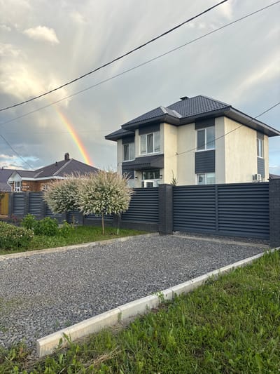 The image shows a modern two-story house with a light-colored stucco exterior and dark gray roof and accents, located in the village of Nagaevo, Russia. A dark gray metal fence encloses the property, with a gravel driveway leading to it. In the background, a rainbow is visible in a cloudy sky, arching over a neighboring house. Two ornamental trees with white blooms are planted in front of the fence. The foreground features a patch of green grass and a concrete curb. Power lines are visible in the sky.