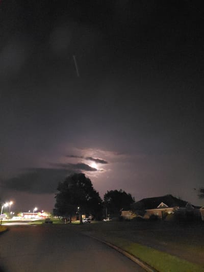 This is a nighttime image taken from a residential street in Nicholasville, Kentucky, United States of America. The sky is dark and cloudy, with a faint light, possibly from the moon or distant lightning, visible behind the clouds. The foreground shows a paved road curving to the left, bordered by a grassy verge. On the right side of the road, there are houses with illuminated windows and manicured lawns. On the left side, there are streetlights illuminating a commercial area with buildings that appear to be shops or businesses, and a parking lot. Several trees are scattered throughout the scene, silhouetted against the night sky. There are no people visible in the image, but there are a few parked cars. The overall atmosphere is quiet and subdued, typical of a residential area at night.
