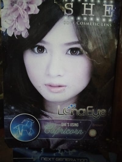 This image depicts a physical advertisement for cosmetic contact lenses, observed in the city of Keo Kouang, Laos. The primary subject is a young East Asian woman with dark hair, a light complexion, and prominent light brown/hazel eyes, indicating the use of cosmetic lenses. Her hair is styled with bangs and a light pink floral accessory is visible on the upper left side of her head. She wears light pink lipstick and maintains a neutral expression, gazing directly forward.
Textual elements are prominently displayed: "SHE" is at the top right, with "SOFT COSMETIC LENS" underneath. Towards the bottom, "LunaEye" is written with a star motif. Below this, the text "SHE'S USING" precedes "Capricorn" in a stylized font, accompanied by a circular blue logo on the left featuring a white constellation outline resembling a goat (Capricorn). To the right of "Capricorn," a small brown circle graphic and the word "Brown" are present. The phrase "NEXT GENERATION" is partially visible at the very bottom. The background features a dark, starry, cosmic design. The advertisement itself exhibits minor scuffs, dust, and reflections, consistent with a physical poster or product packaging.