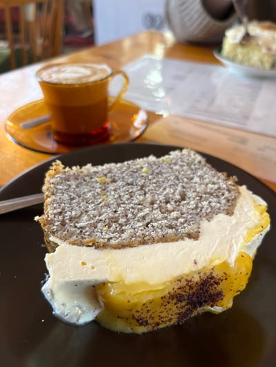 The scene depicts a dessert and beverage presented on a wooden table within an indoor cafe setting in Kaiserslautern, Germany. In the foreground, a multi-layered slice of cake rests on a dark brown plate. The cake comprises an upper grayish-brown layer densely speckled with dark particles, a central thick layer of off-white cream or frosting, and a lower vibrant yellow, semi-translucent layer consistent with fruit curd. This yellow layer partially sags onto the plate, and its bottom edge is dusted with dark brown particulate matter. A small amount of white cream has also dripped onto the plate near the cake's base. A metallic utensil handle is partially visible to the left of the cake. In the blurred midground, an amber-colored glass cup with a handle sits on a matching saucer, containing a beverage topped with white foam arranged in a spiral pattern, indicative of latte art. A small spoon rests on the saucer. The heavily blurred background reveals a light-toned wooden table surface, a folded white paper document (likely a menu), a partial view of another dessert on a plate, and indistinct elements of cafe furniture, including portions of a wooden chair and a woven decorative item. No individuals are visible.
