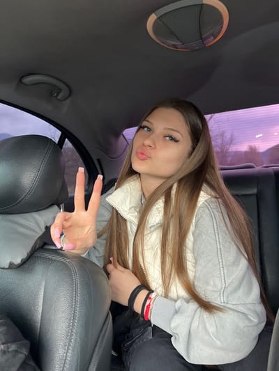 The image displays a young female seated in the rear passenger compartment of a vehicle, located in Cherniche, Bulgaria. The subject, positioned slightly to the right of the frame, has long, straight brown hair and blue eyes. She is looking directly at the camera with her lips puckered in a "kiss" pose. Her left hand is raised to head height, forming a "peace" sign with the index and middle fingers extended; her fingernails are long and feature pink tips. She wears a light-colored (possibly off-white or cream) quilted vest over a grey long-sleeved garment and dark pants, with a visible seam resembling a cargo pocket on her right thigh. Multiple bracelets, including black bands, a red braided string, and a silver-toned bangle, are present on her right wrist. The vehicle's interior includes a dark headliner, an overhead grab handle on the left, and a circular overhead light/ventilation unit. Dark, likely leather, car seats are visible, including the back of the front passenger seat on the left. Through the rear window, an outdoor environment with bare tree branches and distant hills is visible under a sky with a purple-pink hue, suggesting either twilight conditions or tinted glass.