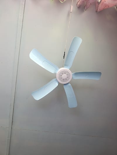 A pale blue, five-bladed ceiling fan is suspended from a light-colored ceiling. The fan's central housing is white with a circular, grooved pattern. A thin, dark string hangs from the fan, connected to a small, light-colored switch or pull cord. The ceiling appears to be constructed of flat, joined panels. In the upper right corner, a portion of decorative fabric, possibly part of a lampshade or wall hanging, with a pink and white paisley pattern is visible. The overall environment suggests an indoor setting, likely a room in a building in Кызыл-Булак, Kyrgyzstan.