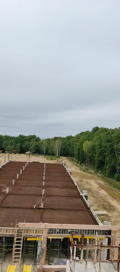 The image depicts a construction site on an overcast day. The foreground shows a large, unfinished concrete slab reinforced with a dense network of rebar. Scaffolding and formwork are visible beneath the slab, indicating ongoing construction work. To the right, a dirt path winds through the site, bordered by a line of white concrete blocks. Beyond the construction area, a dense forest of green trees forms the middle ground. The background is dominated by a pale, cloudy sky. In the distance, a construction crane is faintly visible on the left side of the frame. The scene suggests a large-scale construction project is underway in a semi-rural or industrial area.