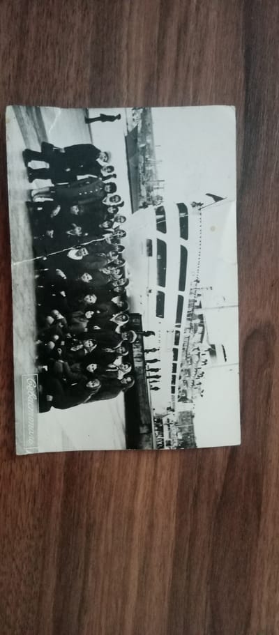 A black and white photograph shows a group of people posing in front of a ship. The individuals are dressed in dark clothing and some are wearing hats. The ship appears to be docked at a port, with city buildings visible in the background. The photograph is oriented vertically, and the text "Соввласт" is printed vertically on the left side. The image is slightly damaged with a vertical crease running through the center. The location in the city of Мариновка, Kazakhstan, is not discernable from the image itself.