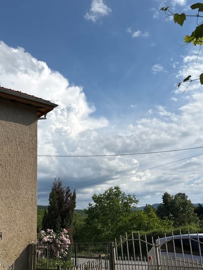 The image captures a view from a residential area in Brusen, Bulgaria, under a partly cloudy sky. In the foreground, the textured stucco wall of a building is visible on the left, with its red-tiled roof extending into the upper left corner. A metal fence runs across the lower part of the image, separating the property from a street. Beyond the fence, a silver car is parked.
The midground features lush greenery, including trees and a cluster of pink flowers in front of a tall, slender dark green tree. In the background, rolling hills are visible, some covered in trees and others appearing as lighter, more distant silhouettes. Overhead, a clear blue sky is punctuated by large, fluffy white cumulus clouds. Tendrils of green leaves and vines hang from the top right corner, suggesting the presence of a nearby plant or tree. Power lines are strung across the scene, contributing to the rural or suburban atmosphere. The overall impression is one of a peaceful, everyday scene in a Bulgarian village.