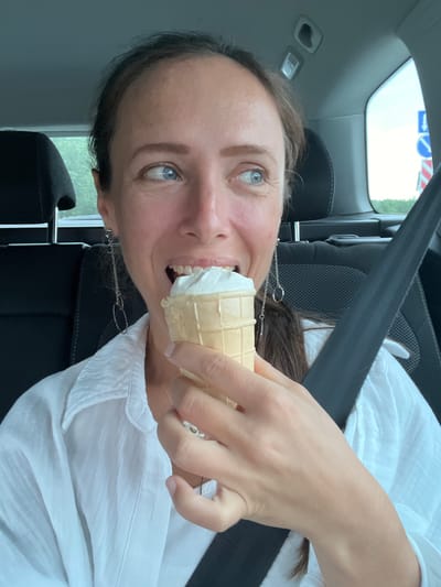 A woman with light brown hair and blue eyes is eating an ice cream cone in a car. She is wearing a white collared shirt and silver earrings. The car appears to be in motion, as indicated by the blurred background of trees and road signs visible through the windows. The woman is looking to her right, with her mouth open and the ice cream cone halfway to her lips. She is wearing a seatbelt. The interior of the car is visible, including the headrests of the seats and the car's ceiling.