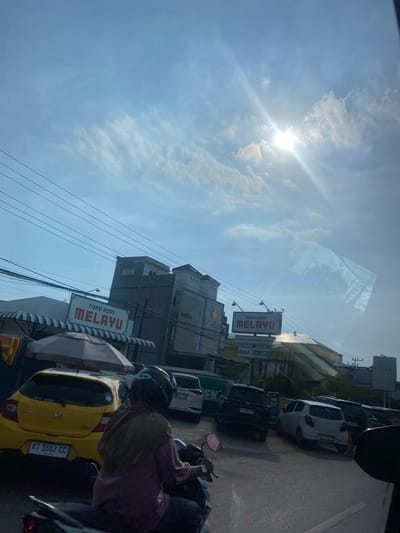 This is a horizontal, eye-level, outdoor shot taken from the perspective of a vehicle. The sky is bright blue with scattered white clouds, and the sun is visible, casting a strong glare. The scene depicts a street in Kertak Hanyar II, Indonesia, with buildings lining the right side of the road.

Several vehicles are present: a yellow car with license plate "KT 1662 CC" is parked on the left, partially obscured by an umbrella structure. Behind it, a black SUV with license plate "DA 1142 PG" is parked, followed by a white car. Further down the road, more cars are parked.

A person is riding a motorcycle in the foreground, facing away from the camera. They are wearing a helmet and a light-colored hijab. The motorcycle has a red taillight.

Signs are visible on the buildings, including a prominent sign that reads "TOKO KOPI MELAYU." Another sign below it indicates "BUKA 24 JAM." Reflections of trees and buildings are visible on the windshield of the vehicle from which the photo is taken. The overall impression is of a busy street scene on a sunny day.