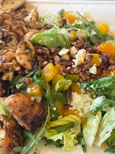 The image shows a close-up of a salad containing grilled chicken pieces, lettuce, arugula, cubed orange segments, pecans, and crumbled cheese. The salad appears to be dressed with a light vinaigrette. While the image does not offer sufficient context to confirm the location as Chula Vista, United States of America.