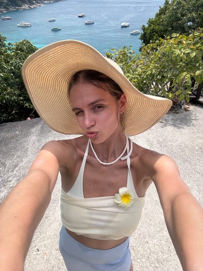 A young adult female takes a selfie outdoors in Lam Kaen, Thailand. She wears a large straw hat, a white top with a frangipani flower, and blue shorts. The background includes a body of water with boats, greenery, and a concrete surface.