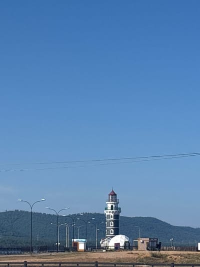 The image shows a black and white striped lighthouse with a red roof, standing on a platform by the water. The lighthouse is located in the city of Turka, Russia, which is situated on the eastern shore of Lake Baikal. The sky is clear blue, and the sun is shining. In the foreground, there is a sandy area with some wooden fences. Behind the lighthouse, there are some buildings, including what appears to be a ticket booth with the word "КАССА" (KASSA) written on it, which means "ticket office" or "cashier" in Russian. Several streetlights are also visible. In the background, there is a lush green hill covered in trees, and the water of Lake Baikal. The scene appears to be a tourist area, possibly a promenade or a recreational spot. There are no people visible in the image.
