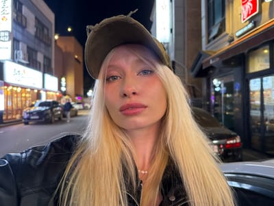 The image displays a close-up selfie of a fair-skinned individual on a street at night in Namyangju-si, South Korea. The subject, centrally framed from the chest up, possesses long, platinum blonde hair, blue eyes, and wears a dark-colored cap, a black leather-style jacket, and a pearl necklace. Pink lip makeup is visible. The individual is looking directly into the camera with a neutral expression.

The background reveals a streetscape illuminated by artificial light. On the left, a multi-story building features several illuminated Korean signs, notably "원조한방삼계탕" (Original Herbal Samgyetang) and "식당" (Restaurant) on a prominent sign. The ground level displays windows with warm interior lighting, indicating a food establishment. A dark-colored SUV is visible in the mid-ground on the street, with blurred figures nearby, suggesting pedestrian presence. On the right, another building with a darker facade and an illuminated sign with red characters is partially visible. The overall environment is an urban setting at night, characterized by architectural structures, street lighting, and vehicles.