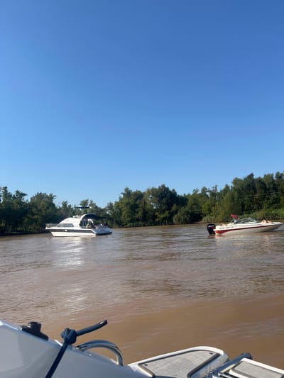 The image depicts a river scene, possibly in San Fernando, Argentina. The water is a muddy brown color. The sky is a clear blue. Trees line the riverbank. Three boats are visible: one white boat is in the foreground, another smaller white boat is further away, and a red and white boat is the furthest away. The photograph appears to be taken from one of the boats.