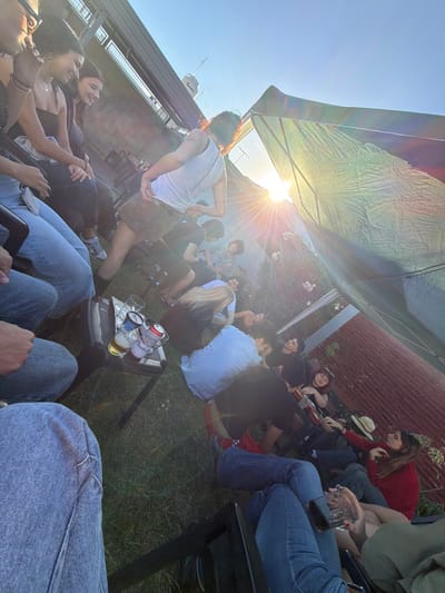 This image depicts an outdoor social gathering in Quilmes Oeste, Argentina, during late afternoon or early evening, characterized by strong, low-angle sunlight originating from the right of the frame. Approximately 15-20 young adults are present, distributed across a grassy area.
In the foreground to the left, the legs and lower torso of a person in light blue jeans are visible, seated on a dark chair. Adjacent to this, a small, dark folding table holds two clear beer glasses (one full, one partially full with amber liquid) and two aluminum cans (one red Budweiser, one silver/white).
To the left, three women are seated; two are smiling and looking towards the center of the group, wearing dark tops. Further into the scene, a person in a white tank top, short brown skirt, and dark boots stands with their back mostly to the camera, appearing to be dancing or moving. This individual has visible tattoos on their upper left arm/shoulder.
Several other individuals are seated or lounging on the grass or low chairs, engaging in conversation. One person in a black beanie and shirt is visible. On the right side of the group, a person is playing an acoustic guitar, while another person next to them wears a red long-sleeved shirt and has a straw cowboy hat resting nearby. One individual in a light-colored shirt is reclining on the ground.
The environment includes a clear blue sky, a large, light-colored fabric canopy or tent structure on the right, intensely backlit by the sun. In the background to the left, a multi-story building with a railing is visible, and a container with a label (possibly "Pernod") is on its rooftop. To the right, a reddish-brown corrugated metal fence forms a boundary, with some green foliage and white flowering plants situated in front of it.