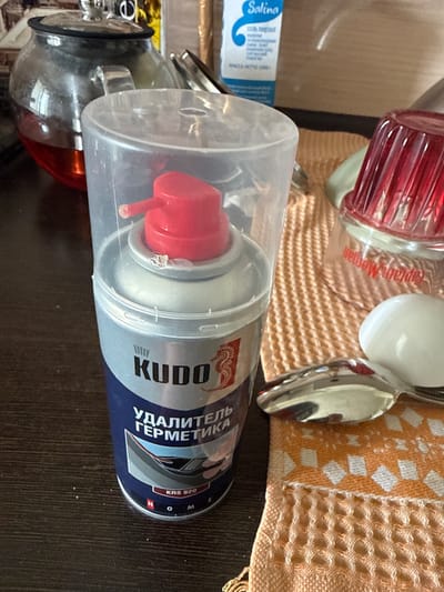 The image shows a close-up of a can of KUDO sealant remover. The can is blue and silver with red lettering and a red nozzle. It is standing upright on a dark wooden surface. To the left of the can, a glass teapot with a red liquid is partially visible. To the right, a patterned orange and white cloth is visible, along with a silver spoon and a white object, possibly a cap or lid. In the background, a box with blue and white lettering that reads "Salina" is also visible. The scene appears to be indoors, possibly a kitchen or workshop. There are no people or animals present in the image. The lighting suggests it is daytime. This image was taken in the city of Romashkovo, Russia.