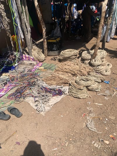 This media file captures an outdoor scene in a marketplace or informal trading area, likely in Nigeria, given the project context. The primary subjects are a large collection of ropes and cords of various thicknesses, colors, and materials, all piled on the ground, some on what appears to be a tarp or fabric. The ropes range from thick, natural fiber ropes bundled together, to thinner, brightly colored synthetic cords, including shades of blue, green, and purple. Some of these are interwoven, suggesting they might be for sale as decorative items or for practical use.
In the background, a rough wooden structure with additional ropes and what looks like fabric or clothing draped over it is visible. A yellow generator is also present in this area. Scattered on the dirt ground are debris, including pieces of plastic and other small objects, indicative of an outdoor, possibly dusty environment. Two dark-colored flip-flops are placed on the ground near the bottom right of the frame, suggesting someone was recently present or is currently in the vicinity. The lighting suggests it is daytime, with clear shadows indicating the sun is at a moderate height. There is no visible text in the image. The overall impression is of a busy, possibly informal trading post where various goods, particularly ropes and cords, are being displayed and sold.