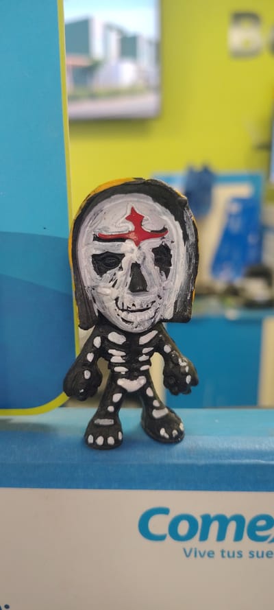 A small, handmade, black-and-white skeleton figure with a red cross on its head lies horizontally on a book with blue and white covers. The book is located on top of a blue and green surface. The location is Chamilpa, Mexico.