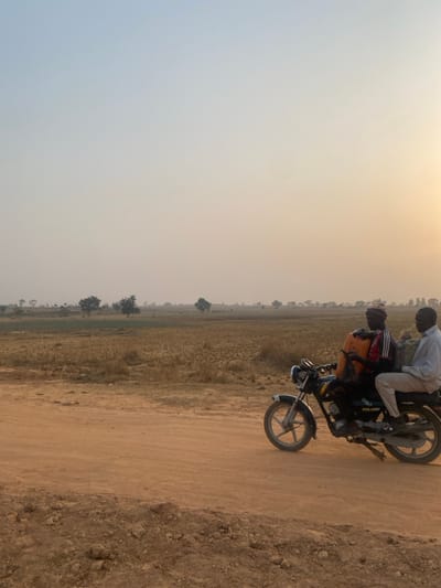 Two people ride a motorcycle on a dusty road. One wears a white shirt, the other a red and black jacket. The road is dirt, bordered by dry grass and scattered trees. The sky is a pale yellow-white, suggesting either dawn or dusk. The overall image composition is somewhat tilted.