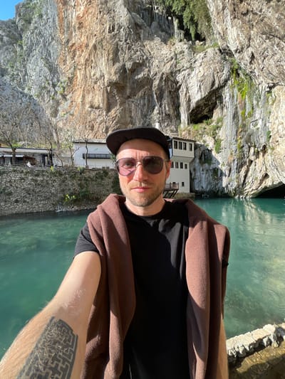 A male individual is positioned in the central foreground, taking a selfie and looking directly at the camera. He wears a black baseball cap, dark aviator-style sunglasses, a black t-shirt, and a brown fleece garment draped over his shoulders. A geometric tattoo with a maze-like pattern is visible on his left forearm. Behind the subject, a body of turquoise-colored water is present, bordered by a low stone wall in the immediate foreground. On the far side of the water, a complex of white, multi-story buildings featuring traditional architecture and dark roofs is situated at the base of a prominent, light-toned limestone cliff. The cliff face, characterized by rugged rock formations and sparse green vegetation, dominates the upper background. A large dark opening, consistent with a cave or grotto, is visible at water level at the base of the cliff. The scene is illuminated by direct sunlight originating from the left. The depicted location is Dračevice, Bosnia and Herzegovina.