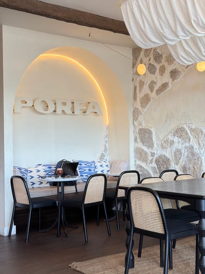 This media file captures an interior scene, possibly a cafe or dining area, with a distinctly modern yet rustic aesthetic. The foreground features a dark wooden floor and a textured rug. A light-colored, arched wall feature dominates the mid-ground, illuminated from behind by a warm, yellow neon light that accentuates the word "PORFA" in large, white, three-dimensional letters.
Behind the neon arch, a table is set with a white marble tabletop, holding a laptop, a backpack, and a glass containing a dark liquid with ice, possibly a drink. The table is surrounded by black-framed chairs with woven cane seats and backs, several of which are visible, suggesting a seating area for patrons. A long, dark wood dining table is also present, lined with more of these chairs.
The background reveals a textured stone wall, partially covered by a creamy white stucco. Hanging from the ceiling are two large, white, pleated fabric lampshades, adding a soft, diffused light to the upper portion of the space. Two small, circular yellow lights are also visible near the ceiling. The overall impression is one of a stylish and inviting establishment, likely open for business given the prepared table setting. The natural light and the warm interior lighting suggest it could be daytime.