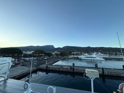 The image captures a wide view of the port area in Papeete, France, during what appears to be early morning or late evening light. In the immediate foreground, the deck of a vessel is visible, featuring a white and orange lifeboat attached to the port side, and a white metal pole on the starboard side with a beige box-like sensor or antenna mounted near its top. Dark water separates this foreground vessel from a concrete pier.

The midground features a marina with dark water, where multiple white yachts are moored. A prominent, multi-deck white superyacht is centrally positioned in the marina. Beyond the pier and marina, a paved road runs parallel to the waterfront. To the left, an extensive parking lot is visible, densely occupied by various cars and trucks. Behind the road and parking area, multiple low-rise buildings with light-colored facades are interspersed among numerous green trees.

The background is dominated by a range of dark green, mountainous terrain, with the peaks on the right catching a subtle, warm light. The sky above is clear, displaying a gradient from a very light blue near the horizon to a deeper blue overhead. No discernible human figures or specific actions are visible.