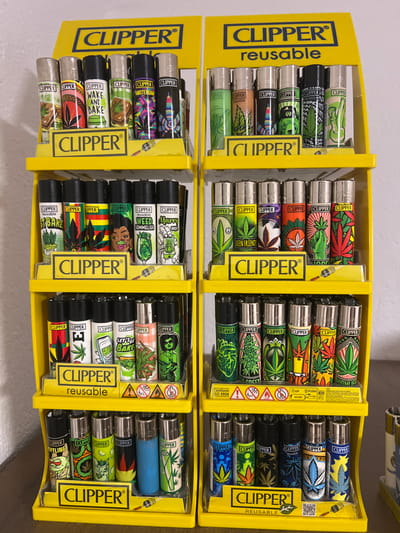 Two bright yellow, multi-tiered display stands, each branded "CLIPPER® reusable," are arranged side-by-side. Each stand features four tiers, displaying rows of approximately six to eight Clipper lighters per tier, oriented vertically. The lighters present a diverse collection of colorful designs, with a prominent recurring theme of cannabis culture, including illustrations of marijuana leaves, cartoon characters, and text such as "WAKE AND BAKE," "WEED SOMMELIER," "AMSTERDAM," and "LET'S GO BAKE." The displays are positioned against a plain white wall and rest on a dark brown surface. No individuals are present, and no actions or interactions are depicted. This scene is located in Mosomishte, Bulgaria.