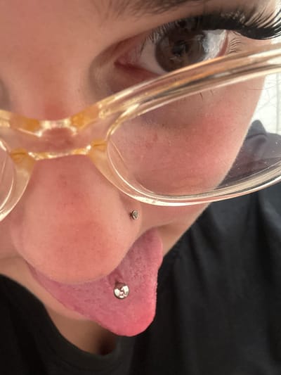 This is a close-up, high-angle, outdoor, low-light, eye-level shot of a person wearing glasses. The person is partially visible, with their mouth and nose in focus. They have a nose piercing on their right nostril and a tongue piercing with a barbell visible on their tongue. The background is blurred, showing a hint of the exterior of a building. The image does not provide any identifiable information about the location in the city of Bad Säckingen, Germany.