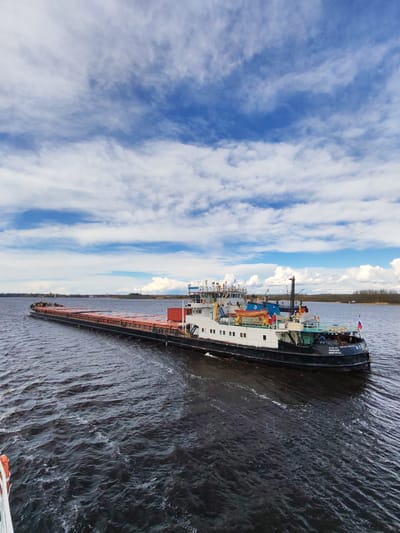 A barge navigates a body of water under a sky filled with scattered clouds. The barge is loaded with cargo, and its structure includes a multi-tiered deckhouse painted in white and black with a red stripe. The stern displays lettering in Cyrillic, identifying the vessel as "VOLGA." The water surface shows choppy conditions. The land is visible in the background. The location is in Russia, near Shestovo.