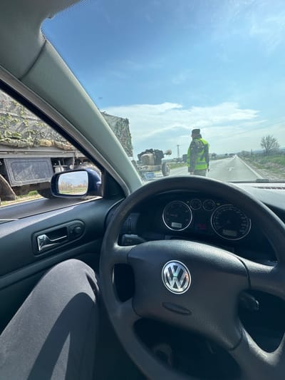 The image depicts a first-person view from the driver's seat of a Volkswagen vehicle, stopped or moving slowly on an asphalt road. The dashboard displays a time of 10:36, an outside temperature of 13.0°C, and an odometer reading of 311,228 kilometers. The driver's left leg, clad in dark pants, is visible.

To the left of the vehicle, a large military truck, camouflaged with netting, is positioned alongside, its rear section visible through the car's window and side mirror. The side mirror reflects a distant view of trees and buildings under a partly cloudy sky.

Straight ahead, a person in a military-style uniform and a high-visibility yellow/green vest stands in the middle of the two-lane road, facing away from the camera. This individual appears to be a checkpoint guard or traffic director. To the immediate left of this person, a camouflaged artillery piece, possibly a field gun or howitzer, is visible, towed on two wheels. The road extends into the distance with white markings, power lines, and sparse vegetation, suggesting a semi-rural setting within or near the city of Manastir, Bulgaria. Other vehicles are faintly visible further down the road under a bright blue sky with scattered clouds.