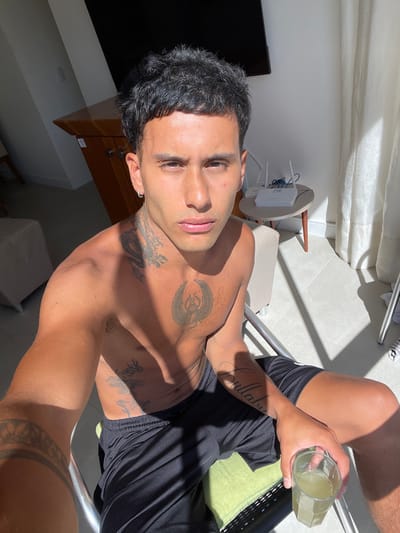 A man with dark hair and light skin poses shirtless, displaying multiple tattoos on his upper body, including one on his neck and another on his forearm. He's holding a glass containing a light-colored liquid. He is wearing black shorts. The scene is sunlit, casting shadows across his body and the surrounding area, which includes a table with a router and a television.
