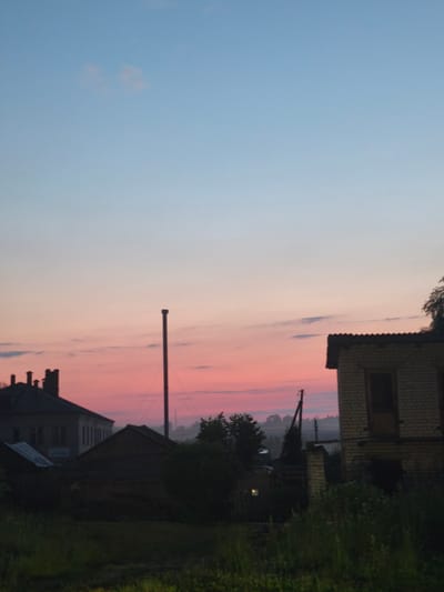 This image depicts a rural sunset scene, likely in the city of Kady, Russia, given the architectural style and vegetation. The sky transitions from a light blue at the top to a vibrant pink and orange gradient near the horizon, indicating the sun is setting. In the foreground, tall green grass and foliage are visible.

Several structures are present. To the left, a multi-story building with a dark roof and multiple windows is silhouetted against the colorful sky. Adjacent to it is a smaller, dark-colored structure with a sloped roof. In the center of the image, a tall, slender pole, possibly a flagpole or antenna, stands out. To the right, a two-story building constructed of light-colored bricks with a corrugated metal roof is prominent. This building has at least one visible doorway and appears to be under construction or in disrepair. Further in the background, hints of trees and other structures can be seen through a light mist. No people are visible in the image.