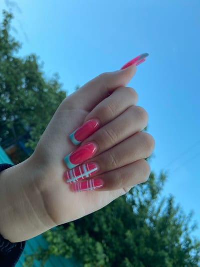A hand with bright pink and turquoise striped and French tip acrylic nails is held up against a clear blue sky. In the background, out of focus, are green trees. The image is taken outdoors. The location of Askarovo, Russia is not depicted in the image.