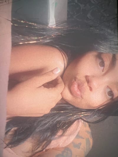 The image is a close-up, vertically oriented photograph of a young woman's face and upper body. She has dark hair, which appears to have some glitter or metallic strands woven into it. Her skin tone is light brown, and she has dark eyes. Her mouth is slightly open, and her left hand is resting on her chin. There is a faint tattoo visible on her shoulder. The lighting is warm and soft, casting a gentle glow on her face. The background is indistinct and appears to be a dark, textured surface. There are some light-colored areas in the background, possibly indicating a wall or fabric. The image appears to be taken indoors, in a casual setting. There is no information in the image to suggest a location in the city of Boone, United States of America.