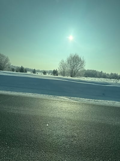 A bright sun shines over a landscape divided by snow and dark pavement. Trees, covered in what appears to be frost, populate the horizon. A thick bank of snow separates the trees from the black asphalt that makes up the foreground. The sun's light reflects off the pavement, creating a shimmer effect. The sky is pale blue, lending a cold aesthetic.