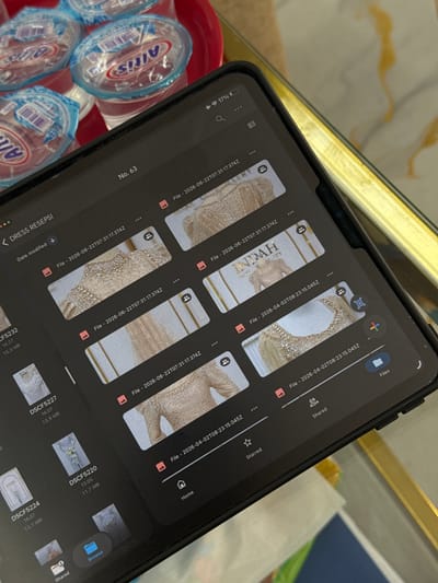 The image shows a tablet device displaying a file management interface, specifically a folder labeled "DRESS RESEPSI". The screen shows multiple image thumbnails, some of which appear to be detailed shots of dresses or clothing with intricate embellishments. File names and dates are visible, indicating a date in 2026. The tablet is placed on a surface that appears to be a table, with several bottled drinks in the background. The lighting suggests an indoor setting. No specific location in Tanjungmeru, Indonesia is indicated by any elements within the image itself.