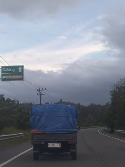 A flatbed truck, covered with a blue tarp, drives on a winding road through a mountainous landscape. The truck's license plate reads "BK 8531 FX". On the left side of the road, a green directional sign indicates "Calang" with an upward arrow and "CRU Sampoiniet" with a leftward arrow. Power lines crisscross the sky, which is filled with dark, heavy clouds, suggesting impending rain or a storm. The time of day appears to be late afternoon or early evening, given the dim lighting. The surrounding environment is lush with trees and vegetation, characteristic of a tropical region like Crakmong, Indonesia. The overall mood is somber and somewhat foreboding due to the stormy weather.