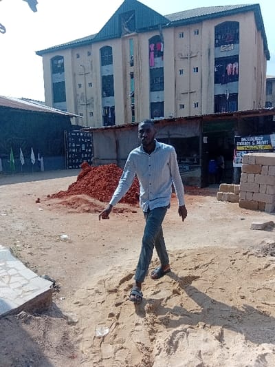 A man stands in a sandy area, seemingly posing for the camera. He wears a light blue shirt and jeans. Behind him, there's a multi-story building with clothes hanging from some of the windows. To the right of the building is a small shop or stall with signage. A pile of red dirt or rubble is in front of the shop. The ground is mostly bare and sandy, with some scattered debris. The image seems to be taken outdoors during daylight.