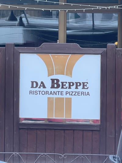 The image displays a sign for an Italian establishment, "DA BEPPE RISTORANTE PIZZERIA," mounted on a dark wooden facade. The sign itself has a white background with a prominent golden or orange abstract design, possibly resembling stylized flames or an abstract chef's hat, with the dark brown text "DA BEPPE" in a larger, bold font, and "RISTORANTE PIZZERIA" in a smaller, sans-serif font below it. The sign is framed by dark brown wood.
To the left of the sign, a large window is visible, reflecting a somewhat blurry scene, possibly showing interior elements or the exterior of an adjacent building. A string of small white lights, likely decorative, hangs vertically along the left edge of the frame, partially obscuring the window. To the right of the sign, a dark, decorative metal railing or gate is partially visible. The overall setting appears to be the exterior of a building, likely a restaurant or pizzeria, consistent with the given location context of Sondrio, Italy. The lighting suggests daytime, possibly overcast, as there are no strong shadows. No people or specific activities are depicted.
