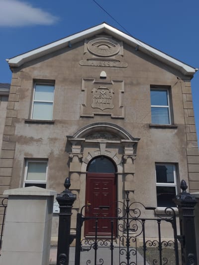 The image shows the front facade of a Masonic Hall located in Magherafelt, United Kingdom. The building is constructed of light-colored stone with some areas showing discoloration and weathering. It has a gable roof with white fascia and soffits. Above the entrance, there is a decorative emblem featuring an eye within a circle and the inscription "A 1899 D". Below this, a shield-like carving displays the Square and Compasses symbol with the words "Masonic Hall". The building has at least two windows on the upper level and two on the lower level, all with white frames. A large, dark red double door is set within an arched entryway, flanked by decorative stone columns. In the foreground, there is a black wrought-iron gate with ornate scrollwork and a stone pillar to the left. The sky is blue with a few white clouds.
